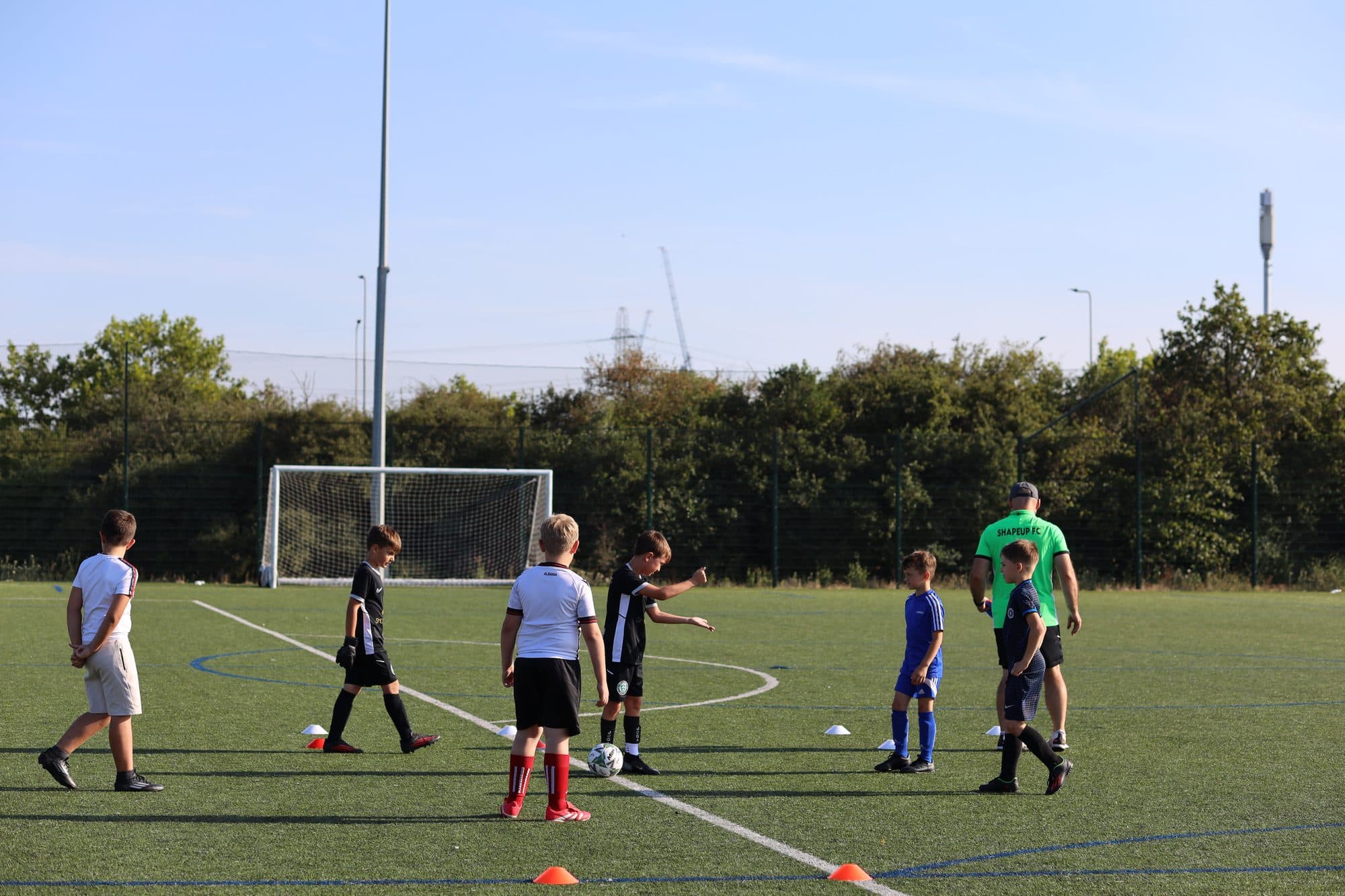 A coach leads a group of young players through a drill on a sunny artificial pitch