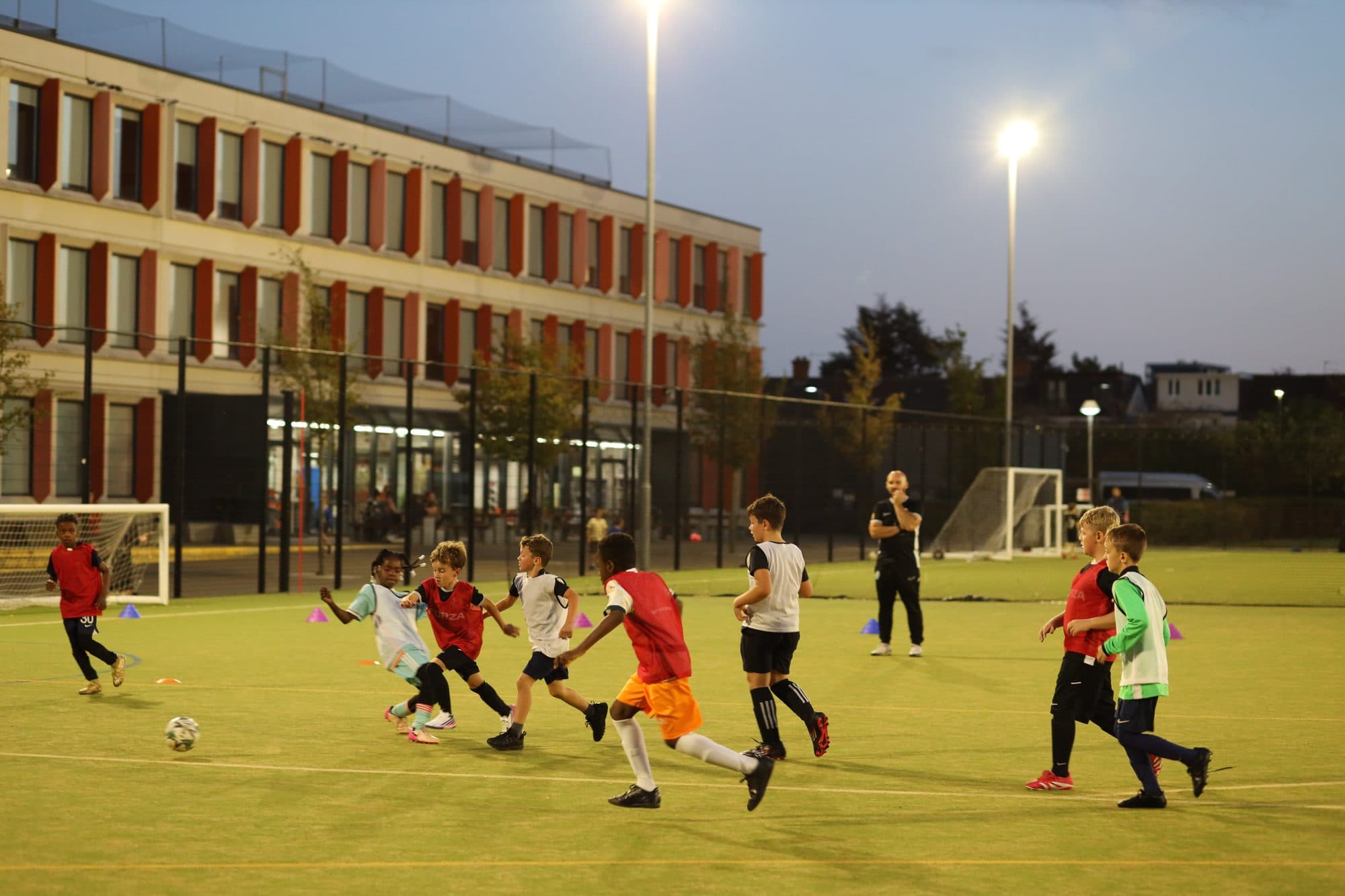 Players chase the ball across a floodlit pitch beside a school building at dusk