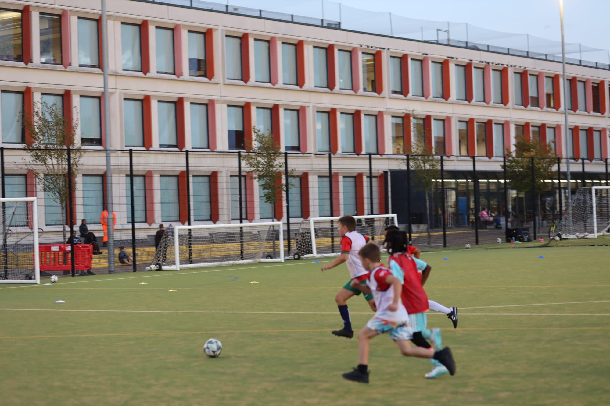 Players sprint after the ball during an evening game on a pitch beside a school building