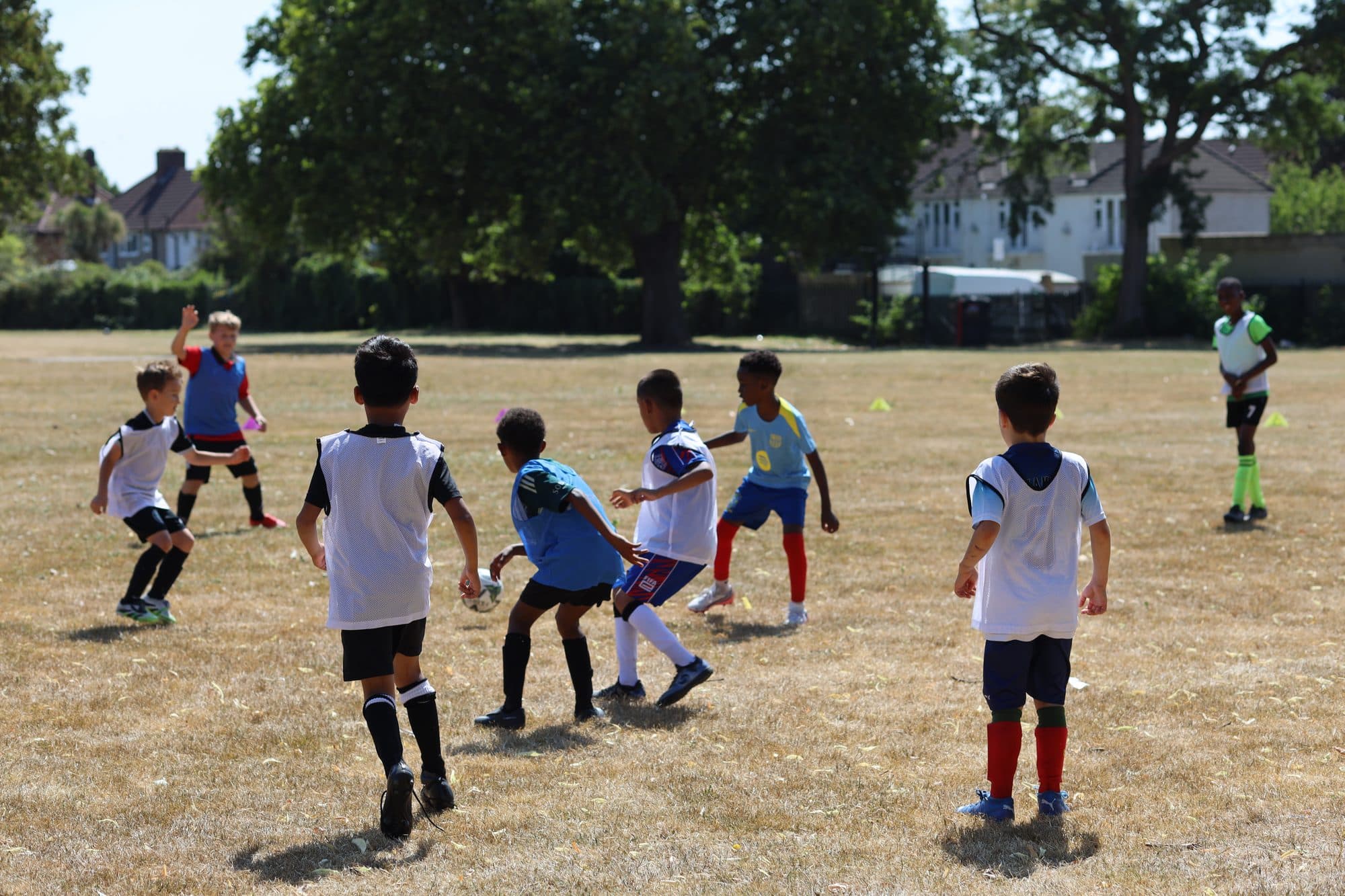 Players in bibs crowd around the ball during a summer game in the park