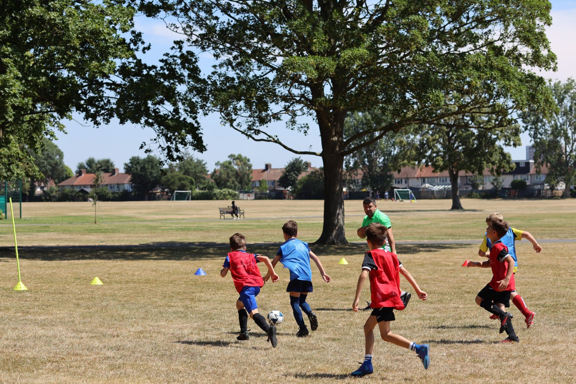 Players in red and blue bibs chase the ball in a game under the trees