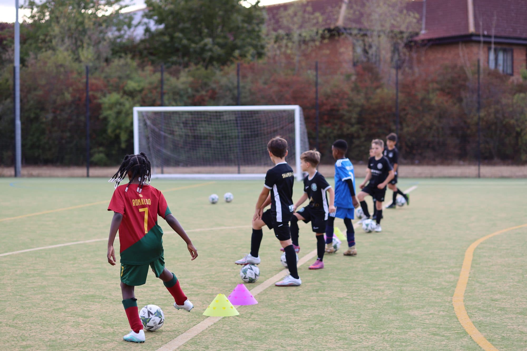 A line of young players dribble footballs between cones on an artificial pitch
