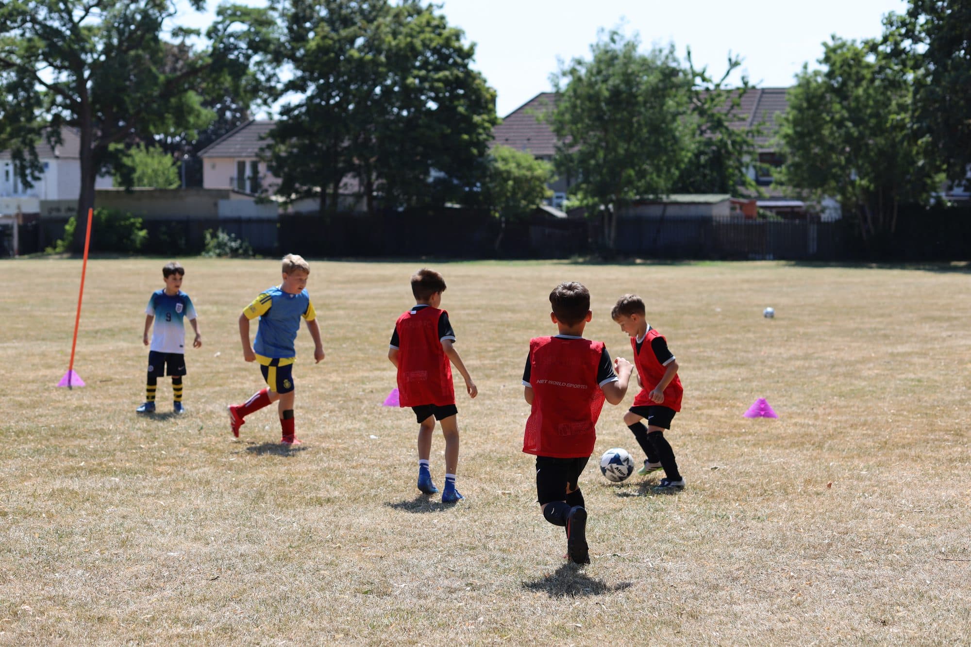 Players in red bibs and blue shirts compete for the ball on dry summer grass