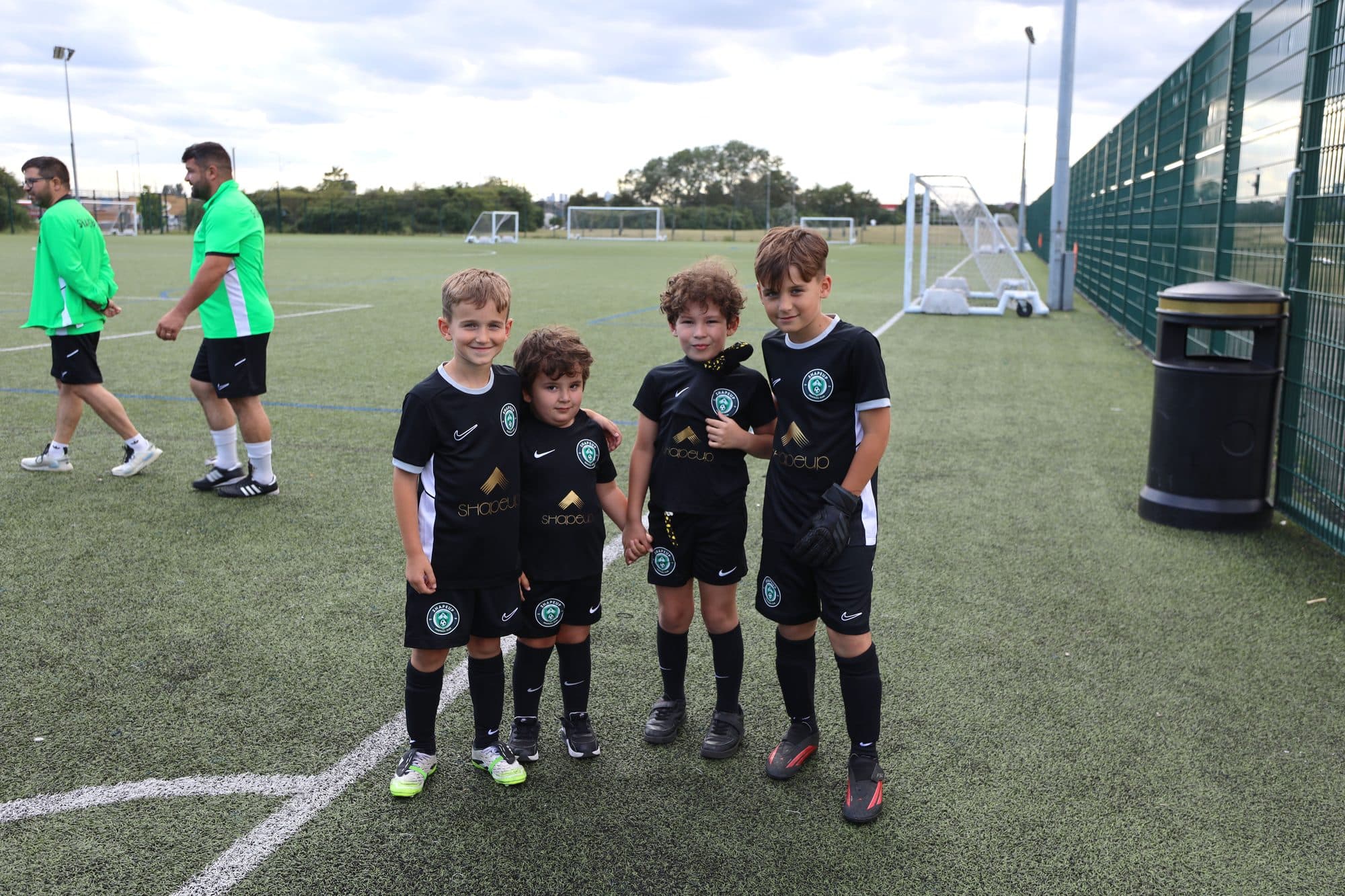 Four young ShapeUp FC players in black club kit smile on an artificial pitch while two coaches in green walk behind them