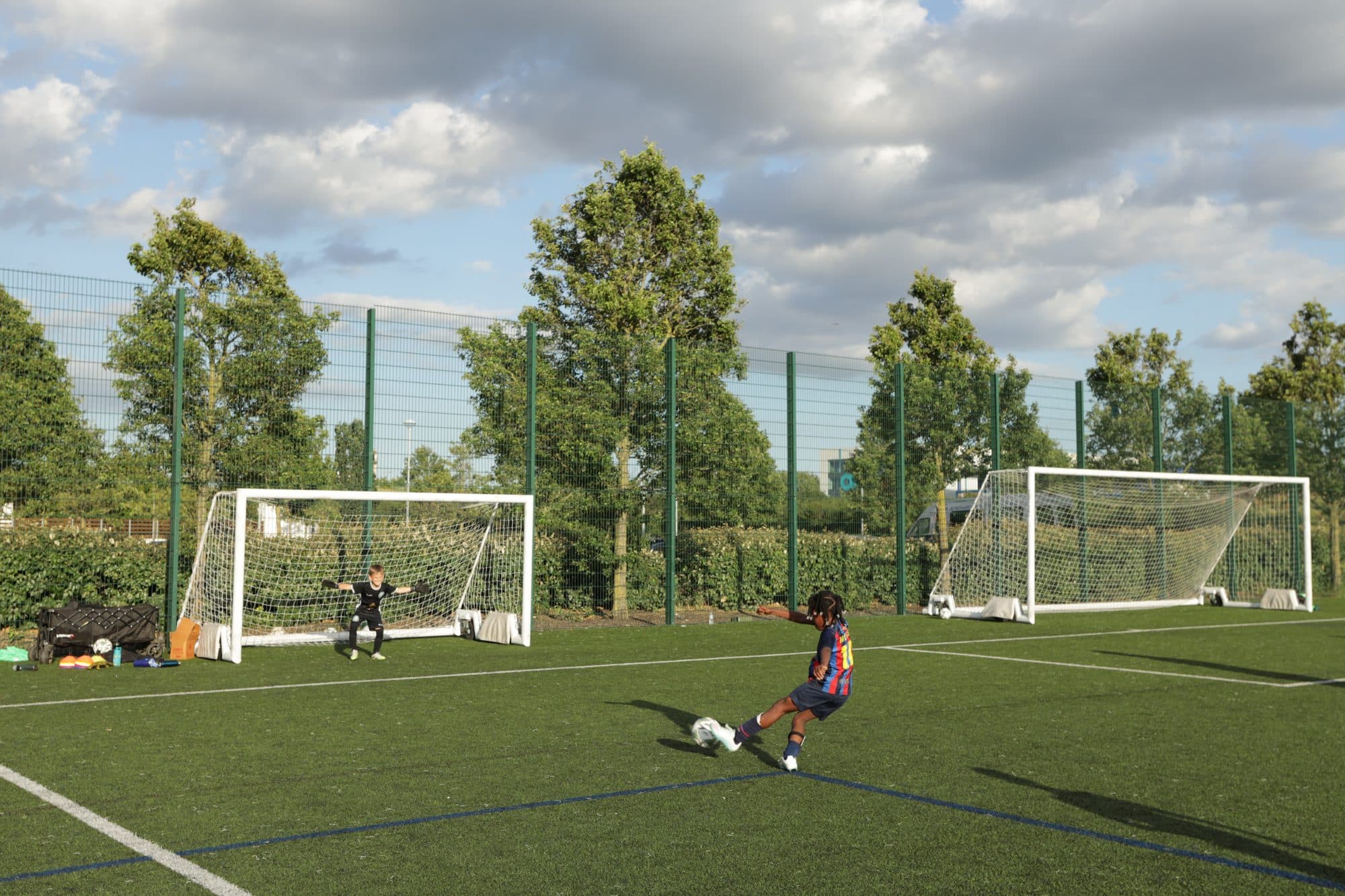 A young player shoots towards goal on an artificial pitch lined with trees