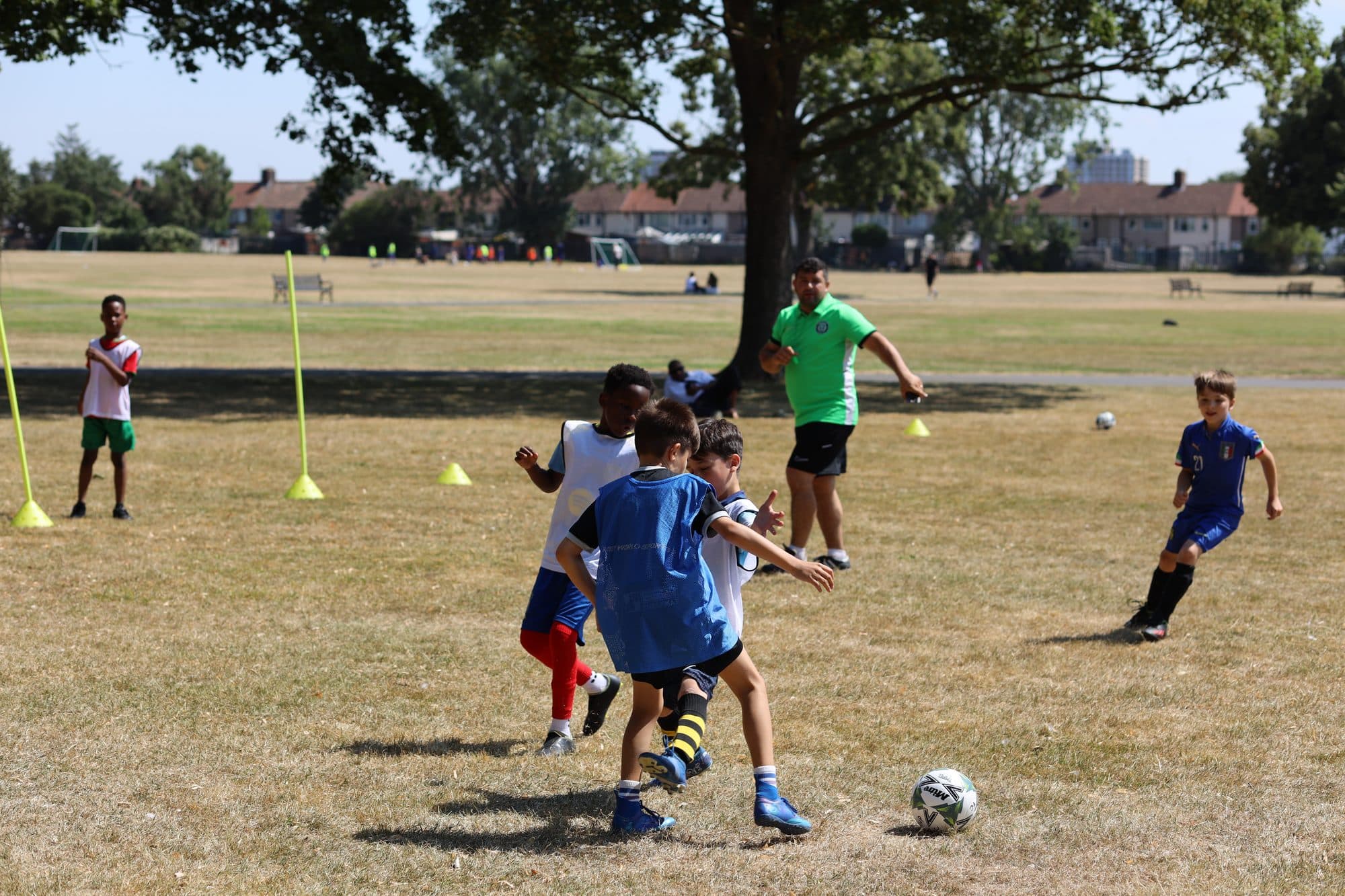 Players chase the ball during a summer game on the grass, with a coach watching on