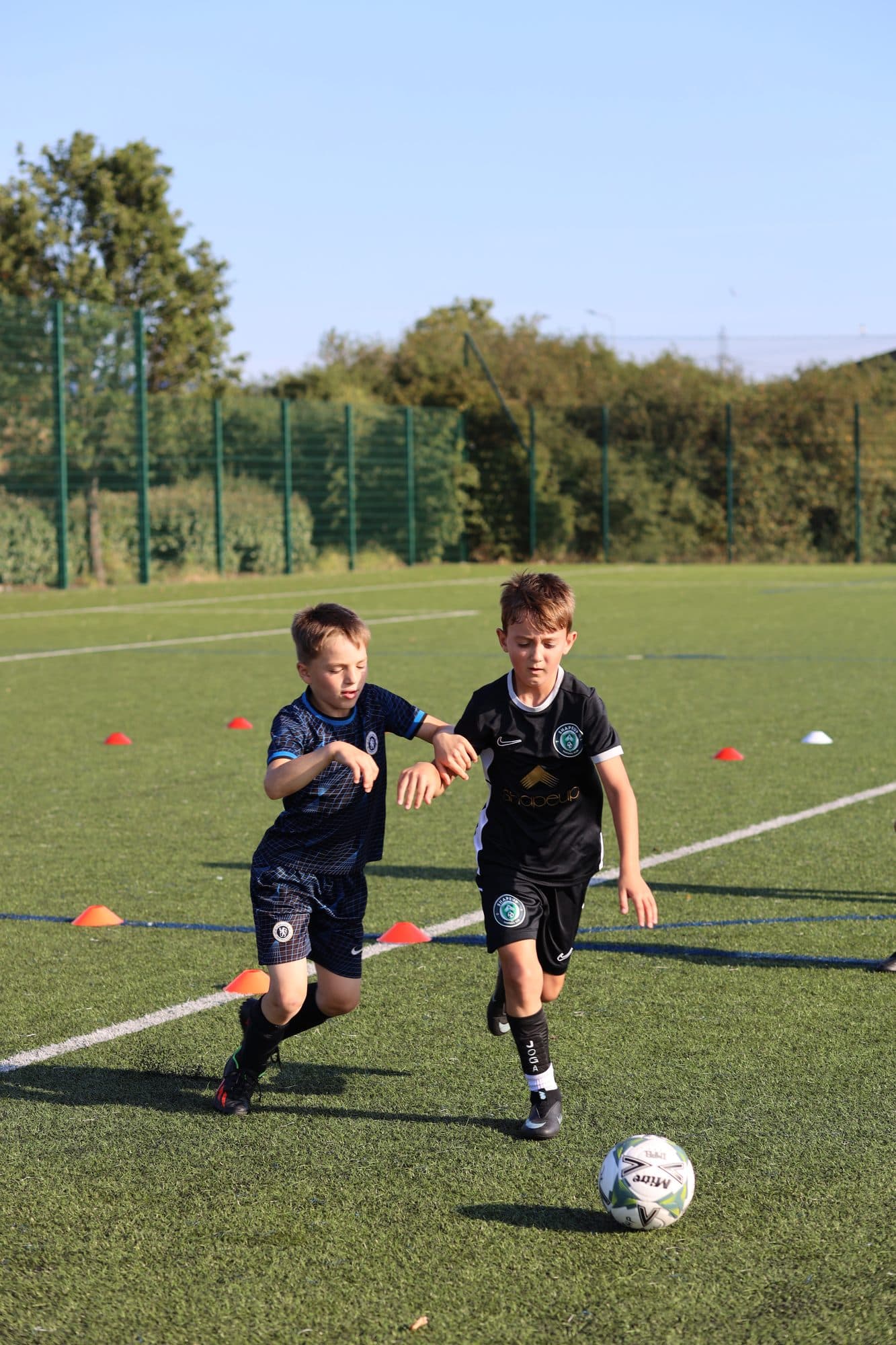 Two young players in matching black kit race for the ball on an artificial pitch
