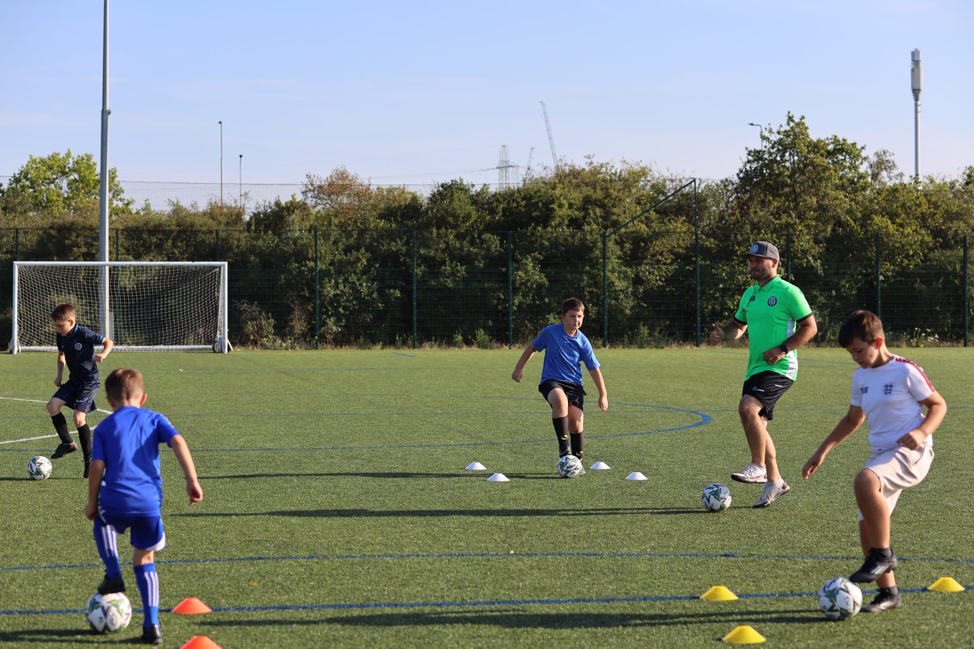 A coach in a green top joins young players in a dribbling drill between cones