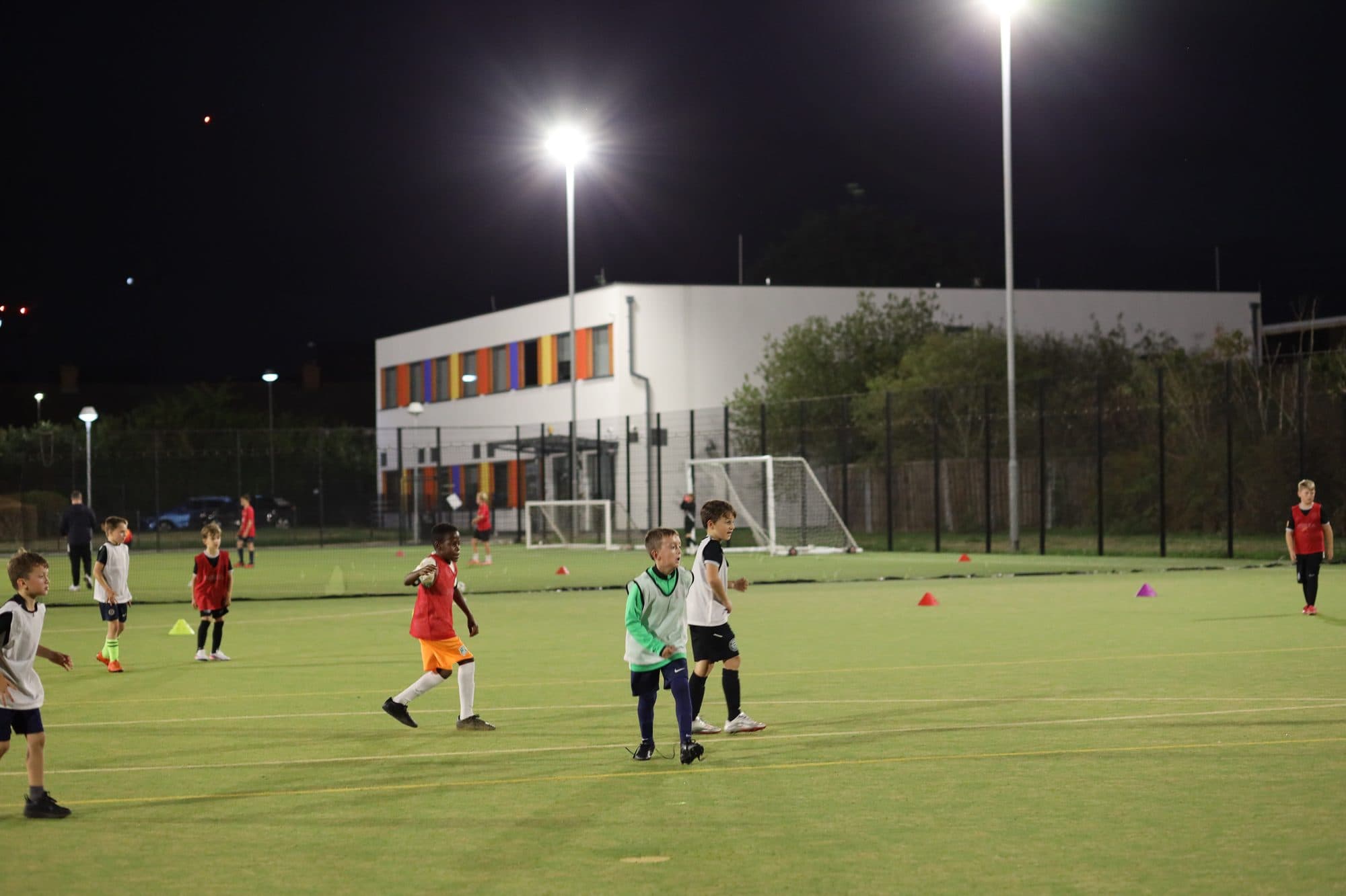 Young players train under bright floodlights on an artificial pitch on a dark evening