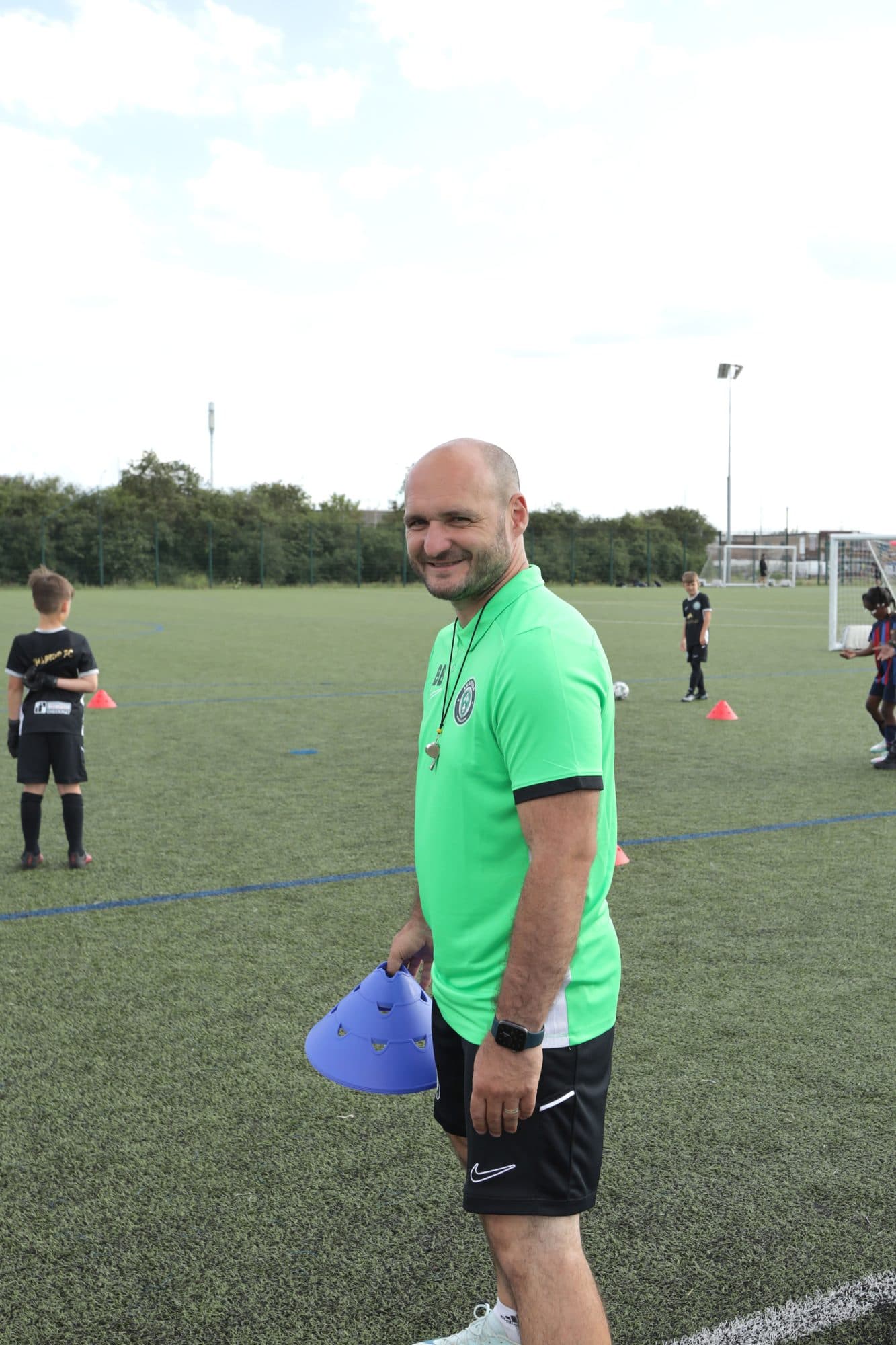 A smiling ShapeUp FC coach in a green top holds training cones on the pitch